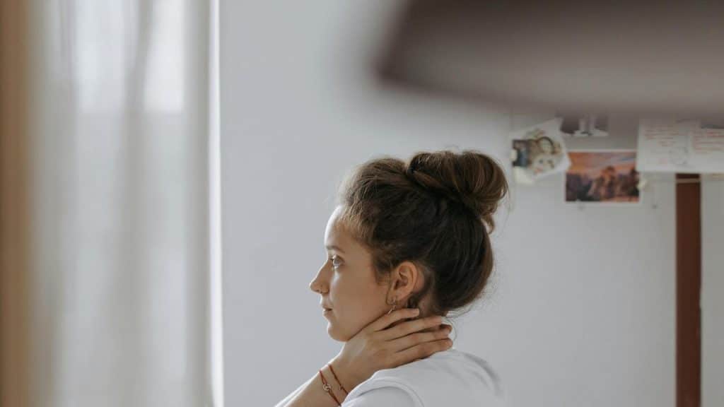 A woman sits indoors with her hand on her neck, looking pensive.