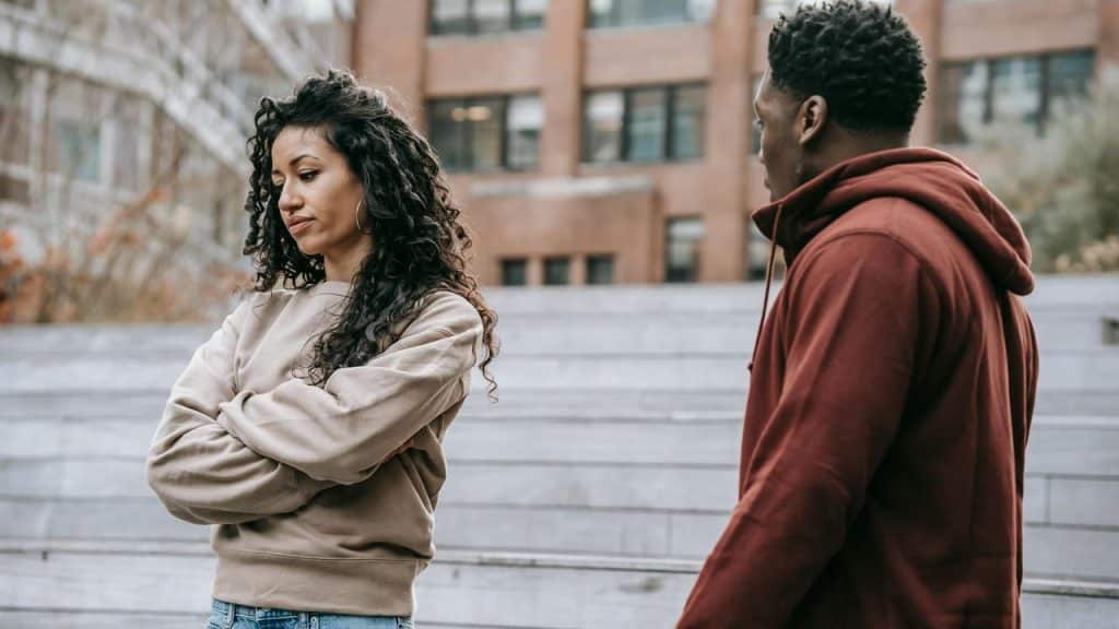 A woman standing with her arms crossed while a man speaks to her outdoors.