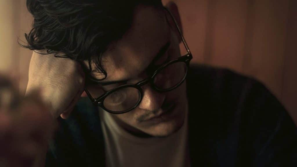 A man wearing glasses is resting his head on his hand with a sad expression.
