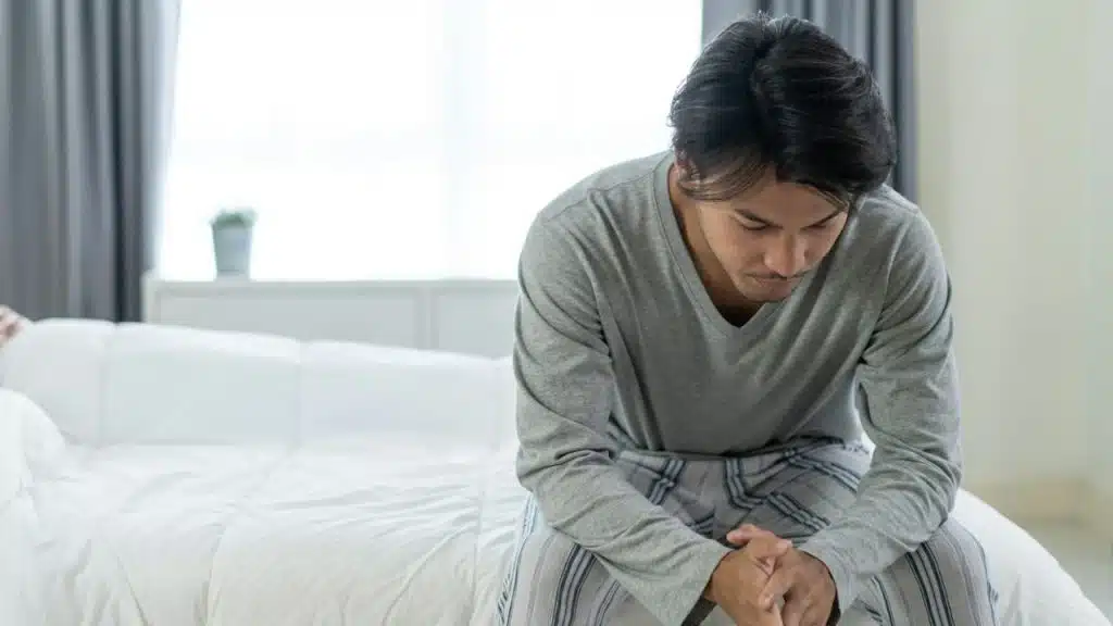 A pensive man in pajamas is sitting on the edge of a bed, looking down.