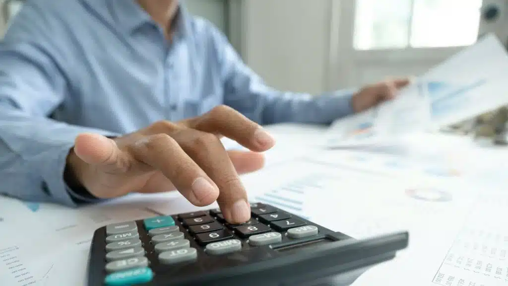 A person's hand pressing buttons on a calculator, with papers and charts visible.