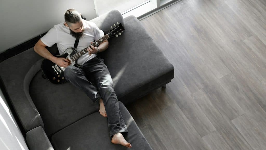 A bearded man playing a guitar while lying on his couch.