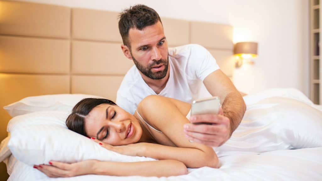 A shocked man leans over a woman, who is smiling with her eyes closed in bed, to look at a smartphone.