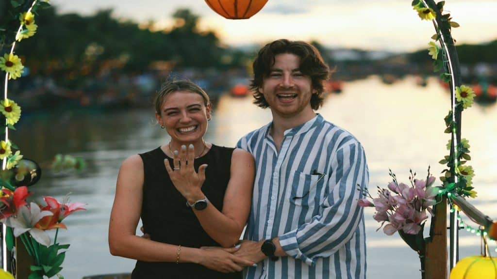 A couple celebrates their engagement under an arch near water.
