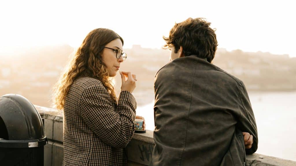 A man and a woman in glasses stand next to each other, looking out from a concrete wall, with the sunset behind them.