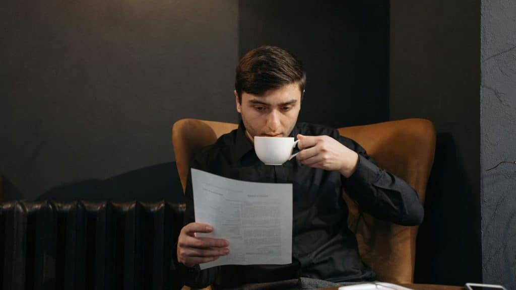A man reading a document while drinking coffee in a cozy chair.