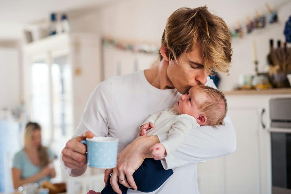 The father is holding the baby in the kitchen while holding a cup of coffee.
