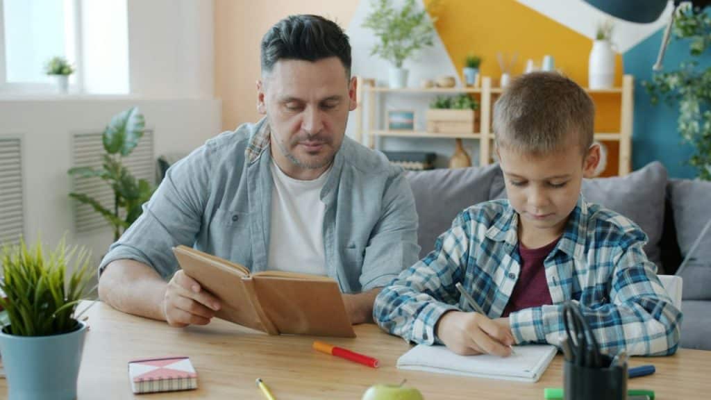 A man is reading a book while a young boy writes in a notebook at a table.