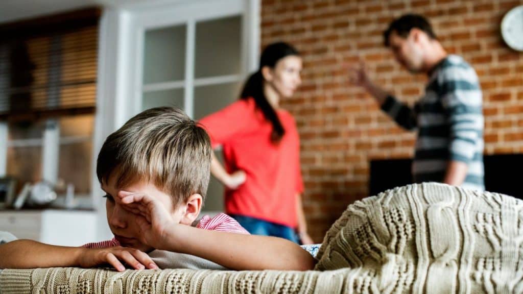A distressed boy covers his eye in the foreground while his parents argue in the background.