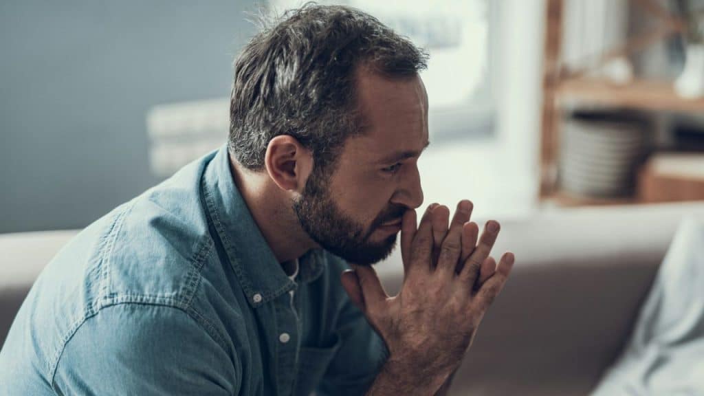 A pensive bearded man in a blue denim shirt sits on a couch, hands clasped near his mouth.