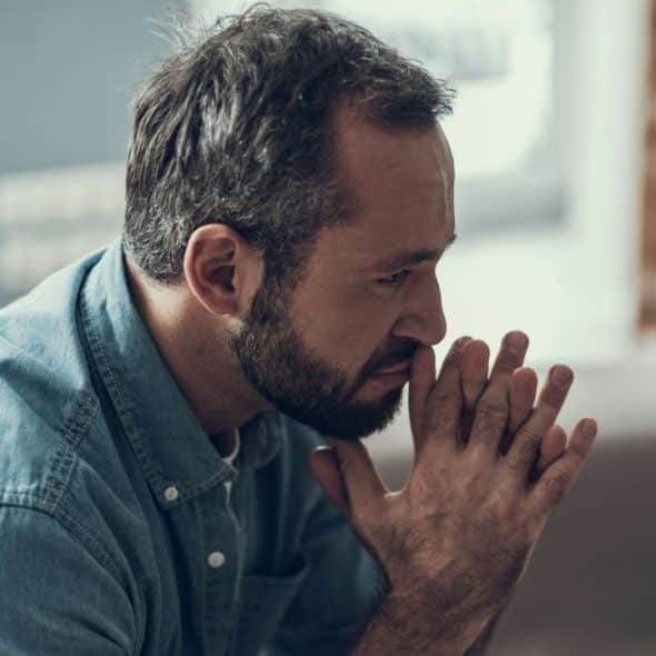 A pensive bearded man in a blue denim shirt sits on a couch, hands clasped near his mouth.