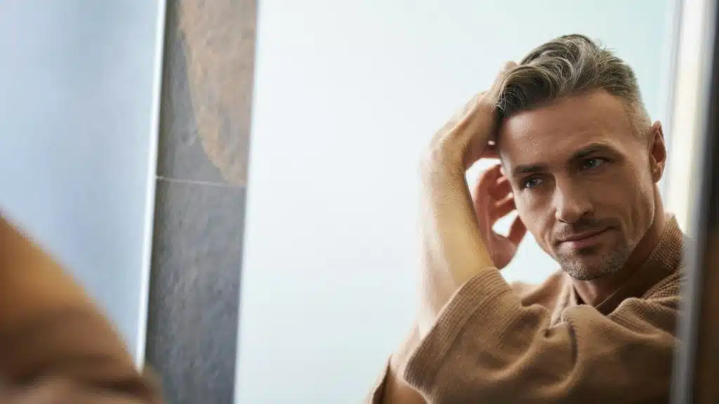 A man in a brown robe looks in a mirror and adjusts his hair with his hand.