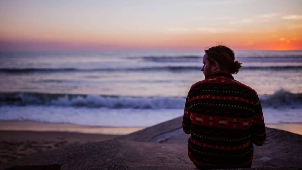 A man in a patterned sweater sits on a beach facing the ocean at sunset.