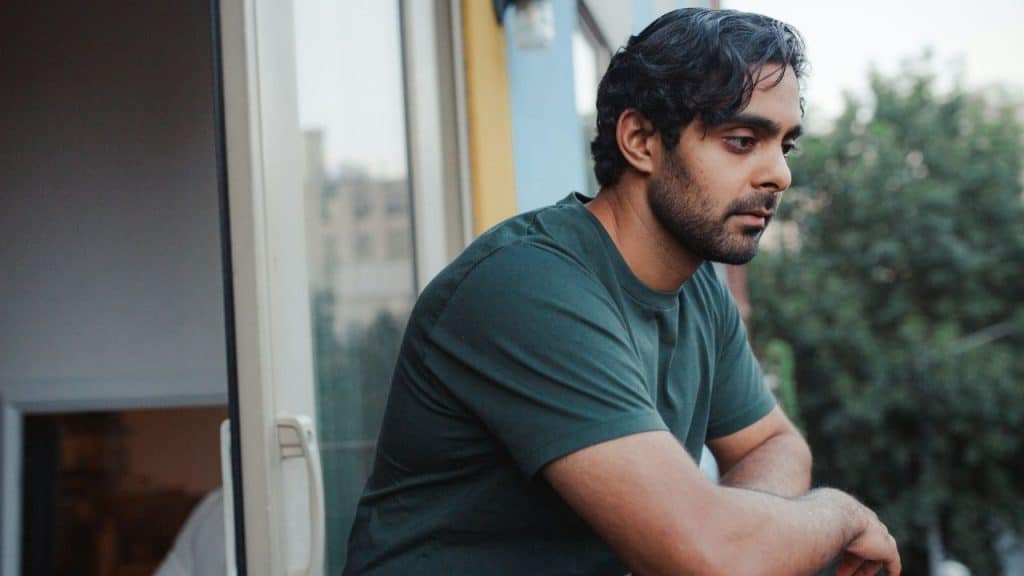 Man in a dark green t-shirt with a beard, leaning on a balcony railing, looking down sadly.