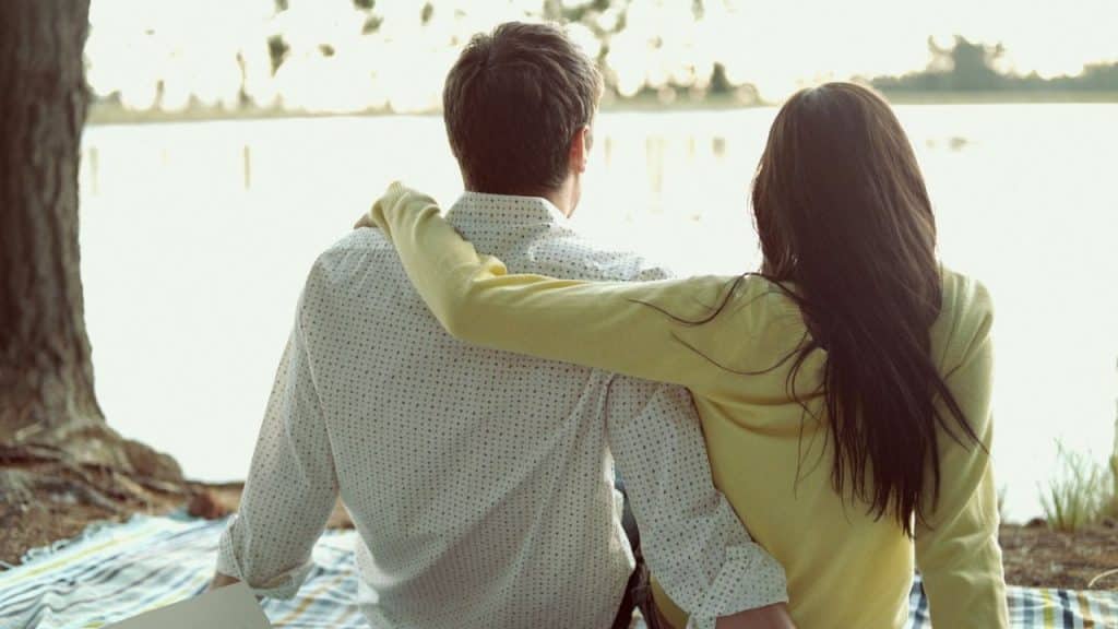 Couple sitting outdoors by the water with their arms around each other, viewed from the back.