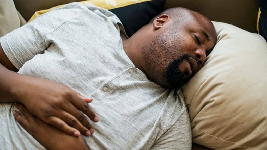 Man with a beard and shaved head sleeping on his side on a couch or bed.