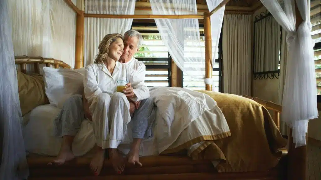 Middle-aged couple in white pajamas sits on a four-poster bed in a tropical room.