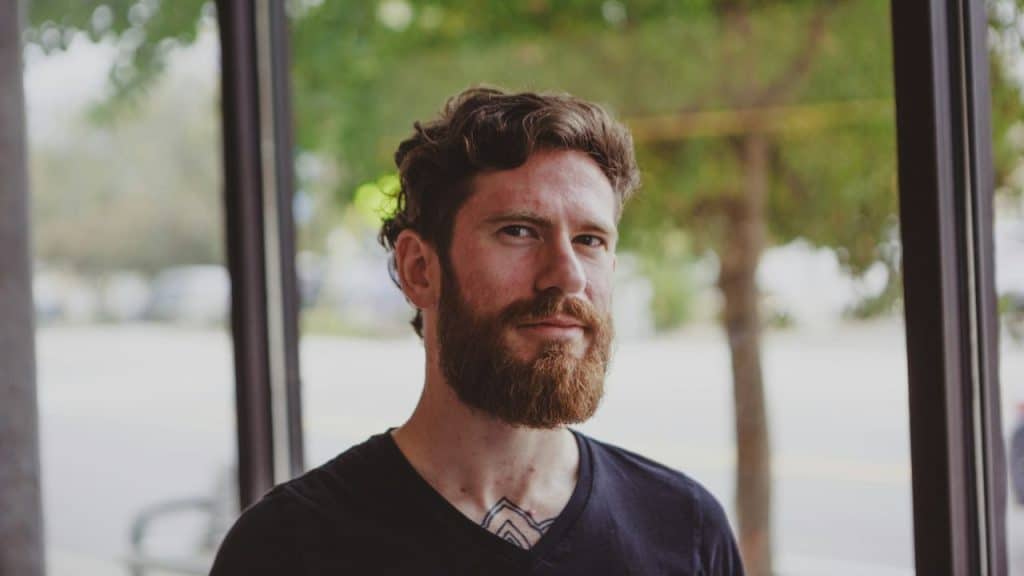 Man with a full beard and curly hair looks forward inside a large window.
