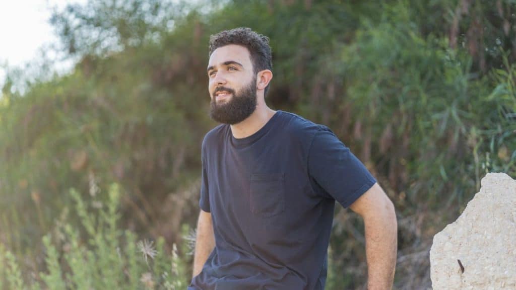 Bearded man in a dark T-shirt looking up and to the left outdoors with green plants.