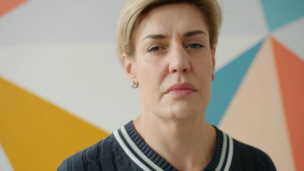 A woman with short light hair and a serious face stands against a geometric background.