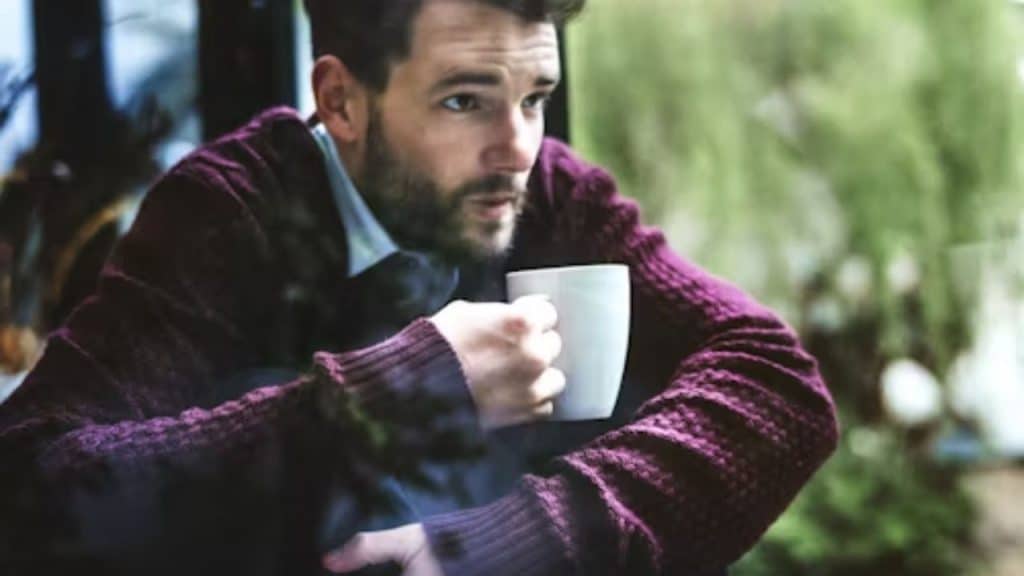 A man looking out a window in deep thought, holding a cup of coffee