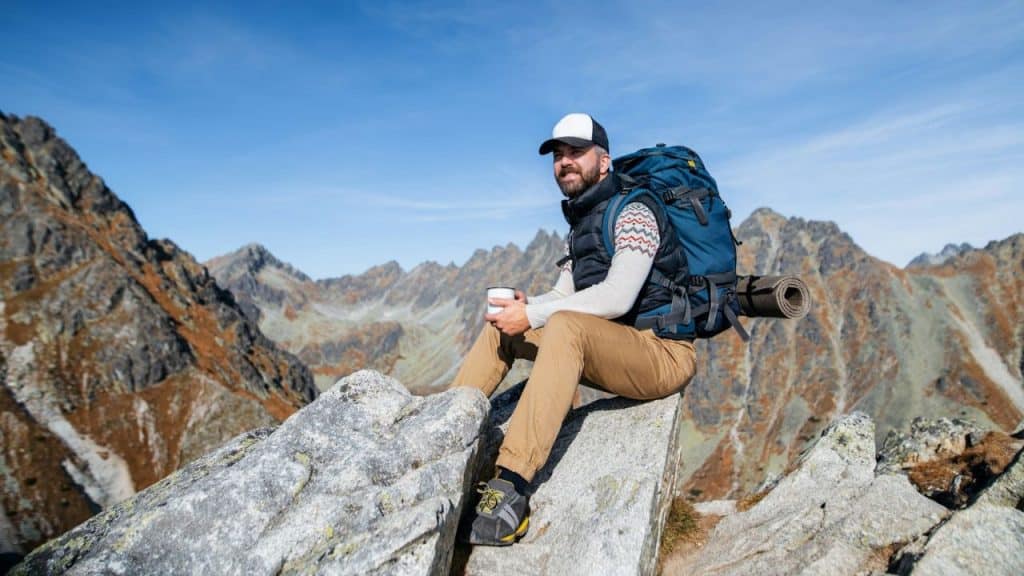 A happy male hiker sits on a rock holding a mug, overlooking mountains under a blue sky.