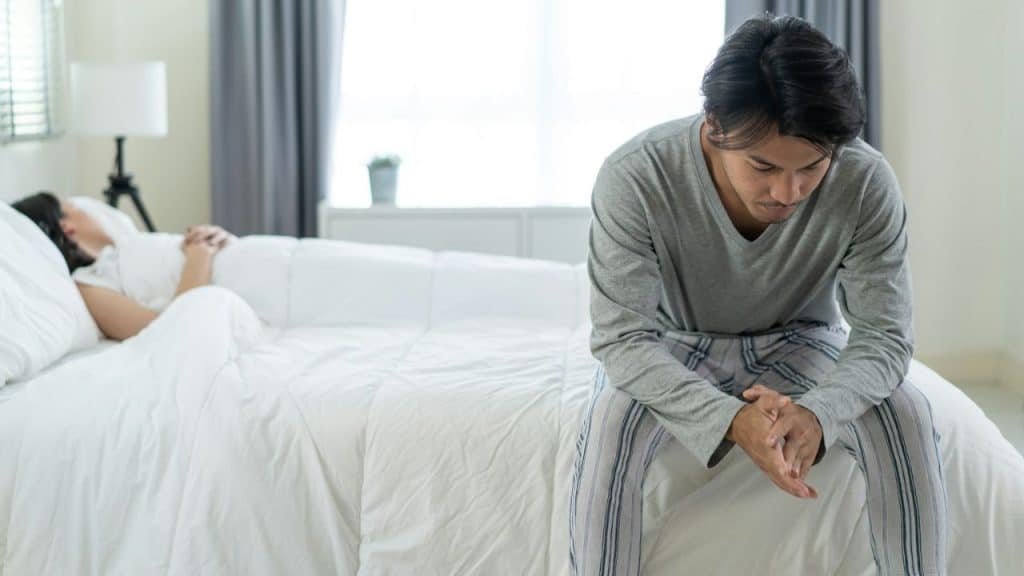 Man in pajamas sitting on the edge of a bed looking down while another person is asleep in the background.