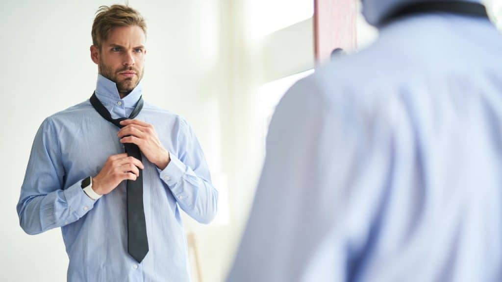 Man in a blue shirt tying a dark tie while looking in a mirror.