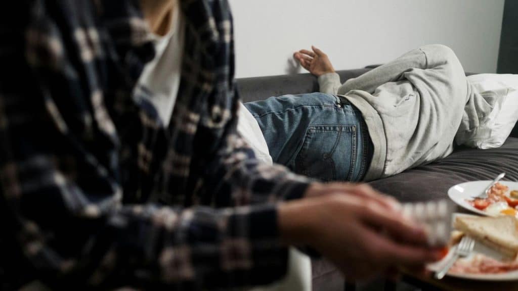 A person lying on a couch with their back turned while another sits nearby holding a drink.