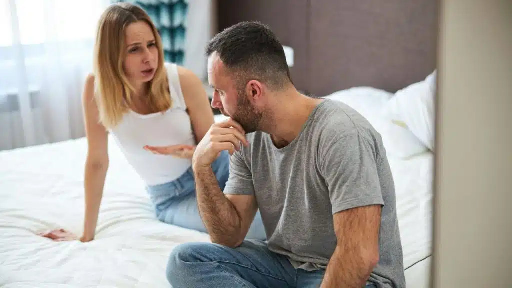 A man sits on a bed with his hand to his mouth while a woman speaks and gestures toward him.