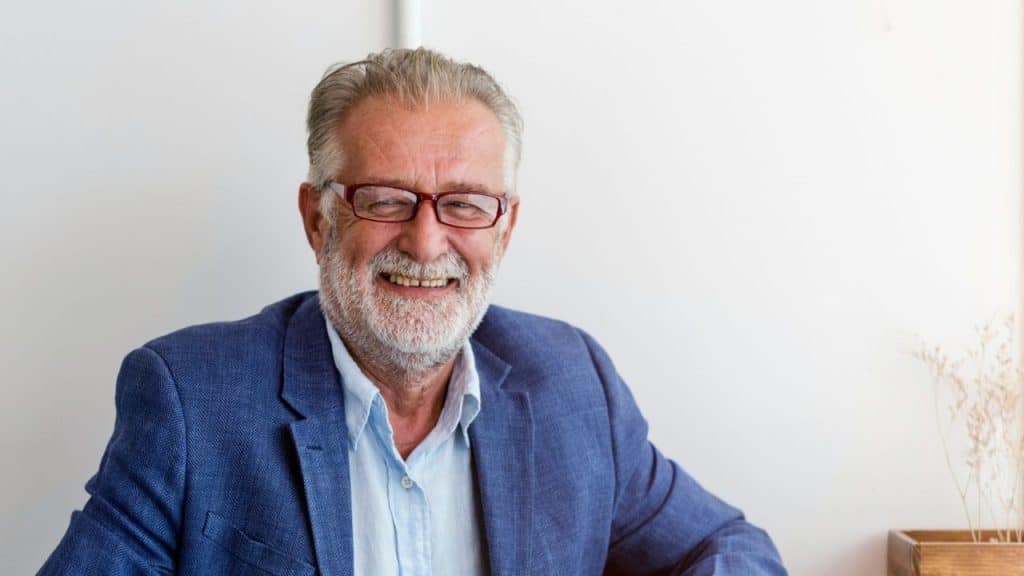 Smiling older man with a white beard, gray hair, and red-rimmed glasses, wearing a blue blazer and light blue shirt.