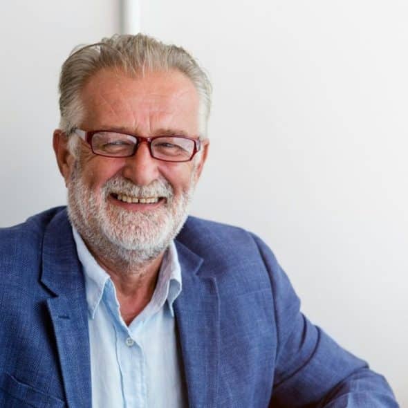 Smiling older man with a white beard, gray hair, and red-rimmed glasses, wearing a blue blazer and light blue shirt.