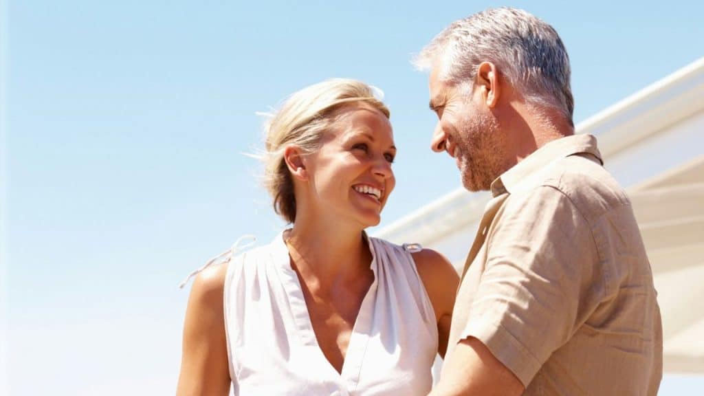 Blonde woman smiling up at a man with gray hair outdoors under a bright sky.