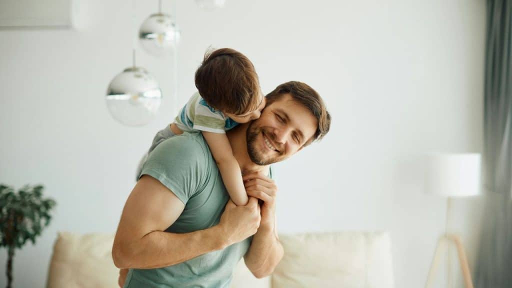 Smiling man giving a piggyback ride to a young child kissing his cheek indoors.