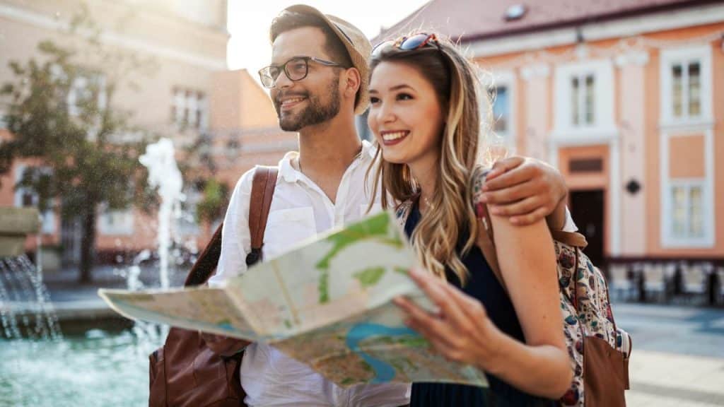 Smiling couple with a backpack and map, standing in a sunny European town square.