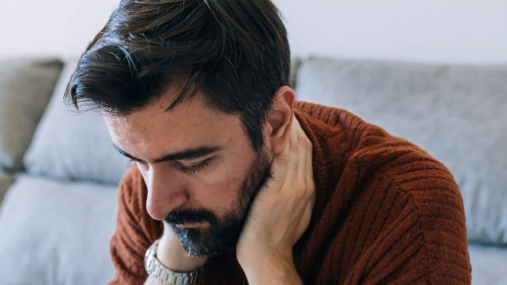 A bearded man in a brown sweater sits looking down, holding the back of his neck.