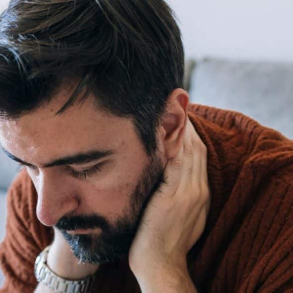 A bearded man in a brown sweater sits looking down, holding the back of his neck.