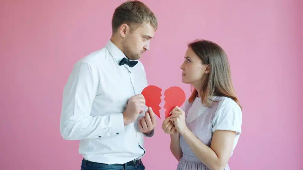 Couple holding broken heart halves on pink background.