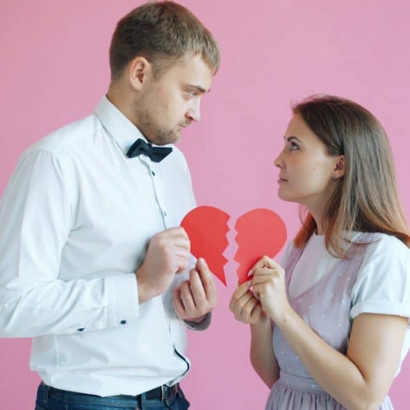 Couple holding broken heart halves on pink background.