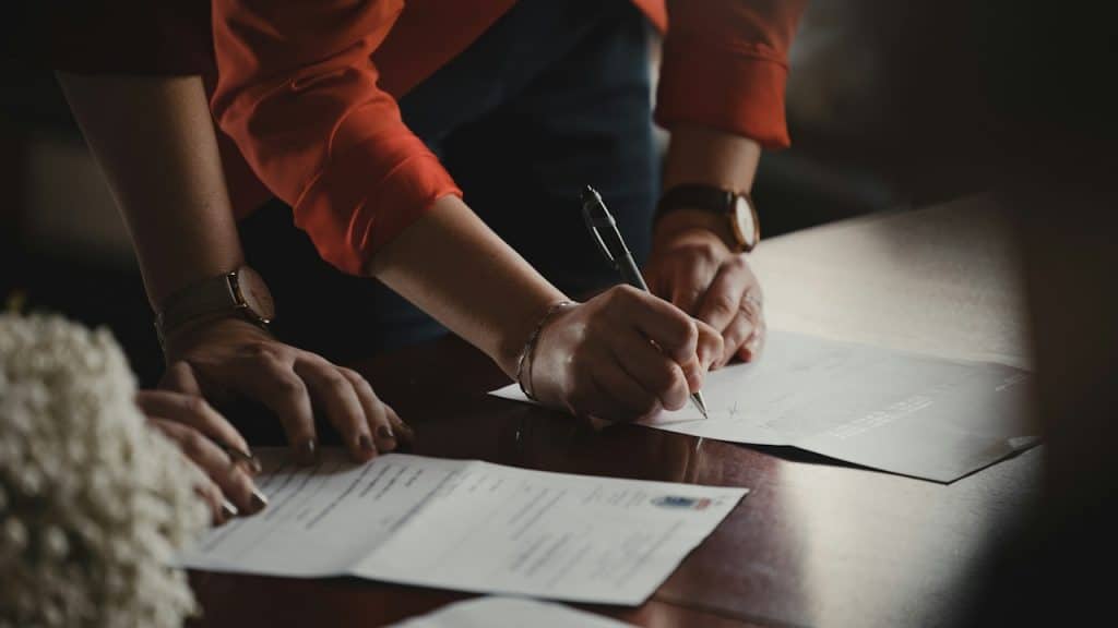 A close-up of two persons signing a document with a pen on a dark wooden table.