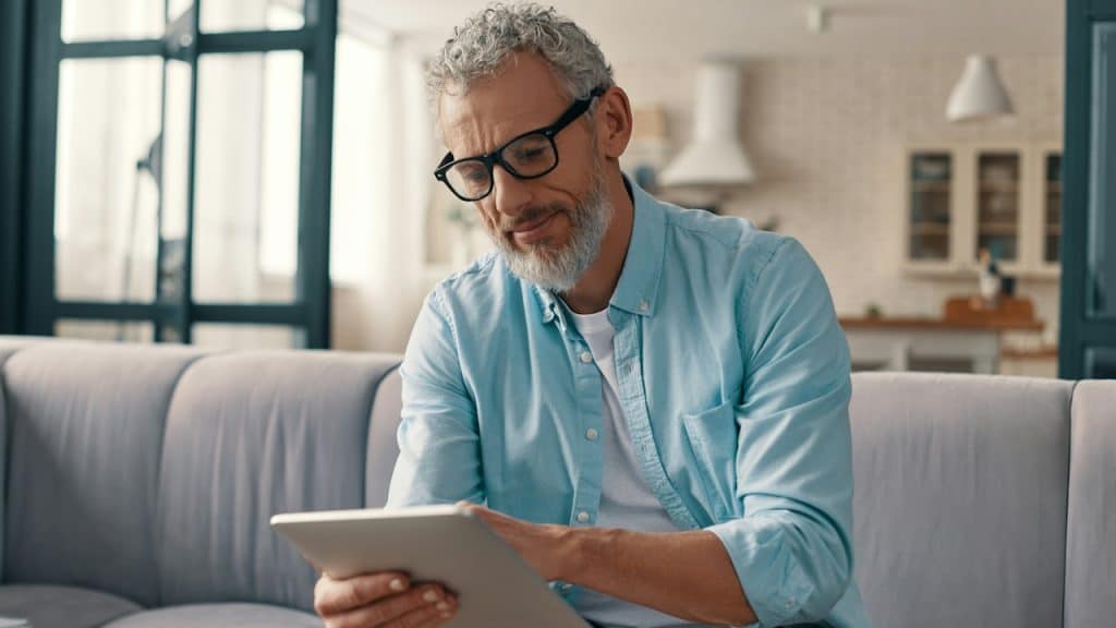 A mature man sitting on a couch and using a tablet.