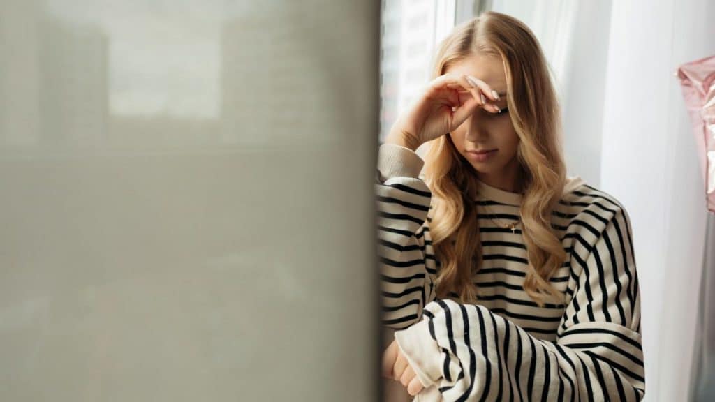 A young blonde woman in a striped sweater sits by a window, covering her eyes with one hand.