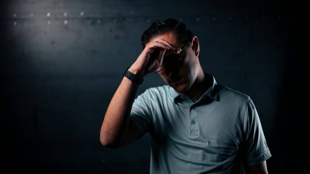 A man in a polo shirt with dark hair and a watch strap looks down, holding his forehead.