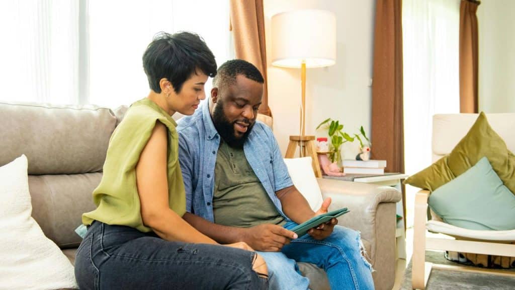 A couple sitting on a couch looking at a tablet together and smiling.