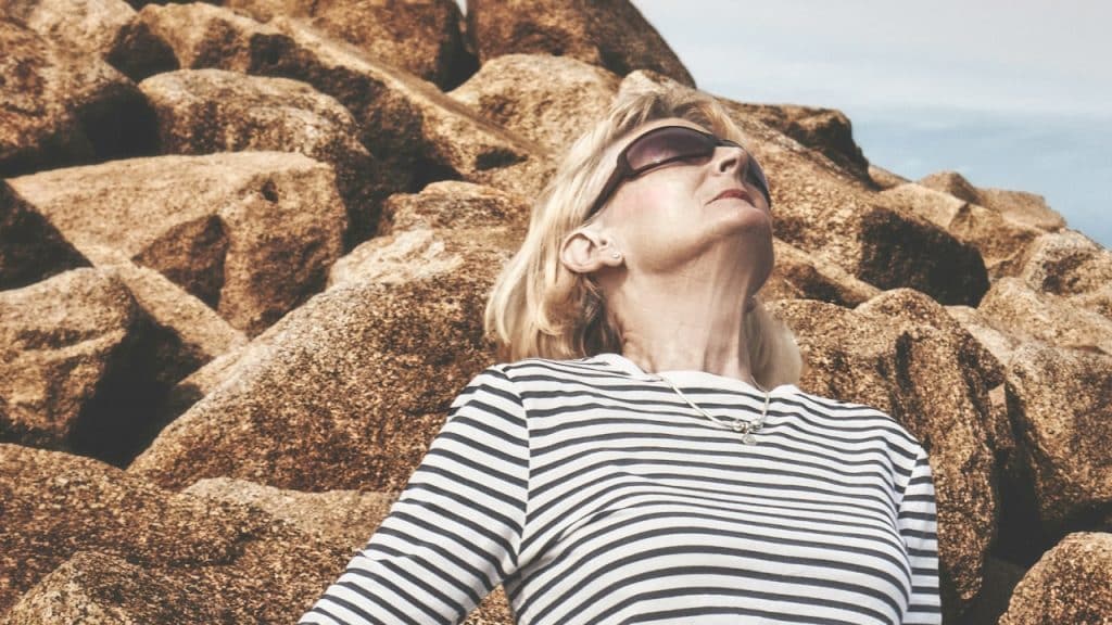 An older woman wearing sunglasses and leaning on rocks.