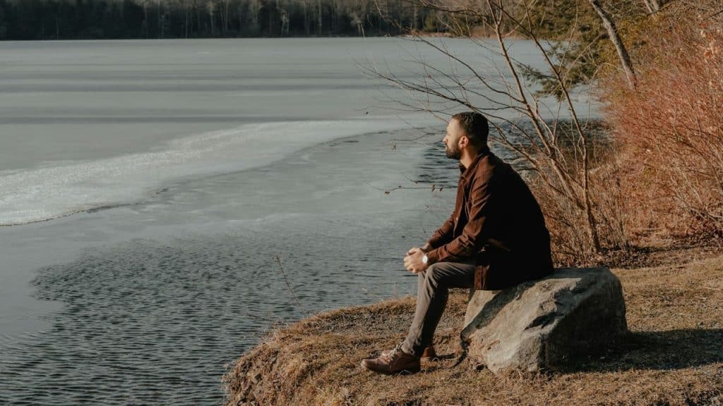 A man in a brown jacket sits on a large rock by a partially frozen lake, looking out over the water.