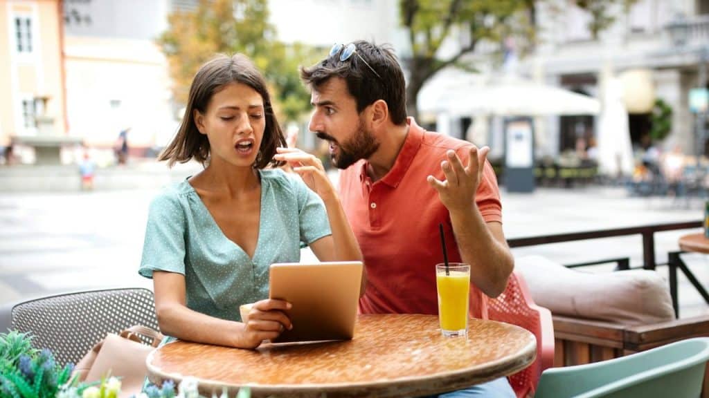 A distressed woman with a tablet sits at an outdoor cafe while a man shouts at her.