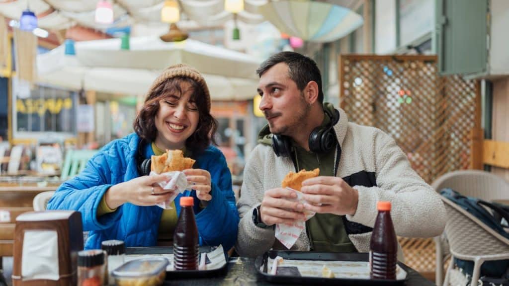 Smiling young couple eating sandwiches at an outdoor cafe with headphones.