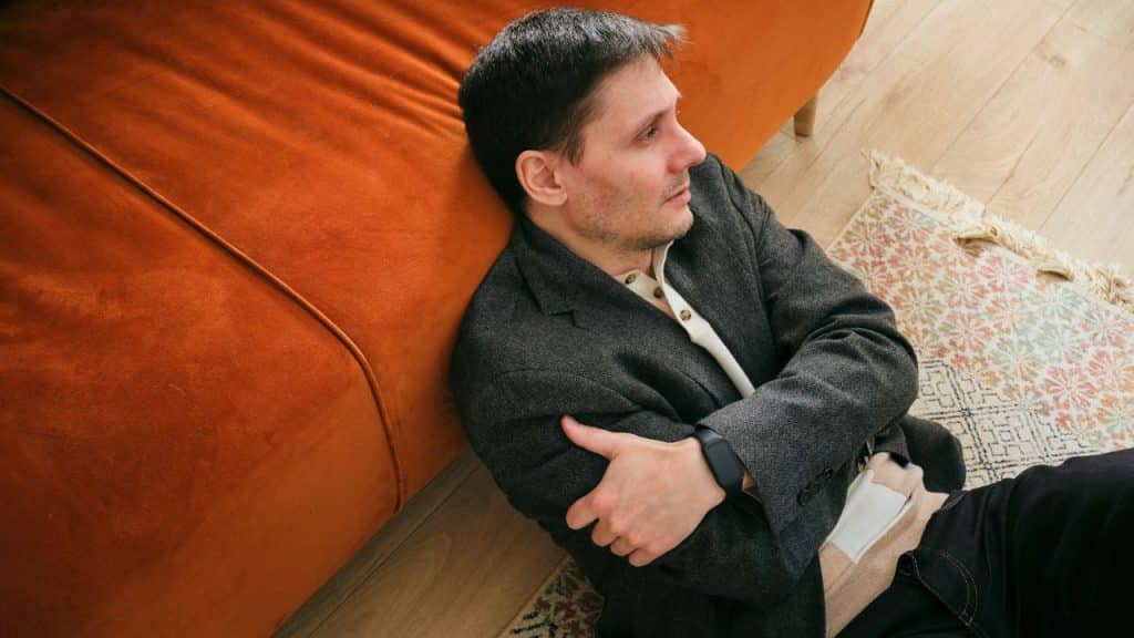 A man in a gray blazer is sitting on a wooden floor, leaning against an orange sofa.