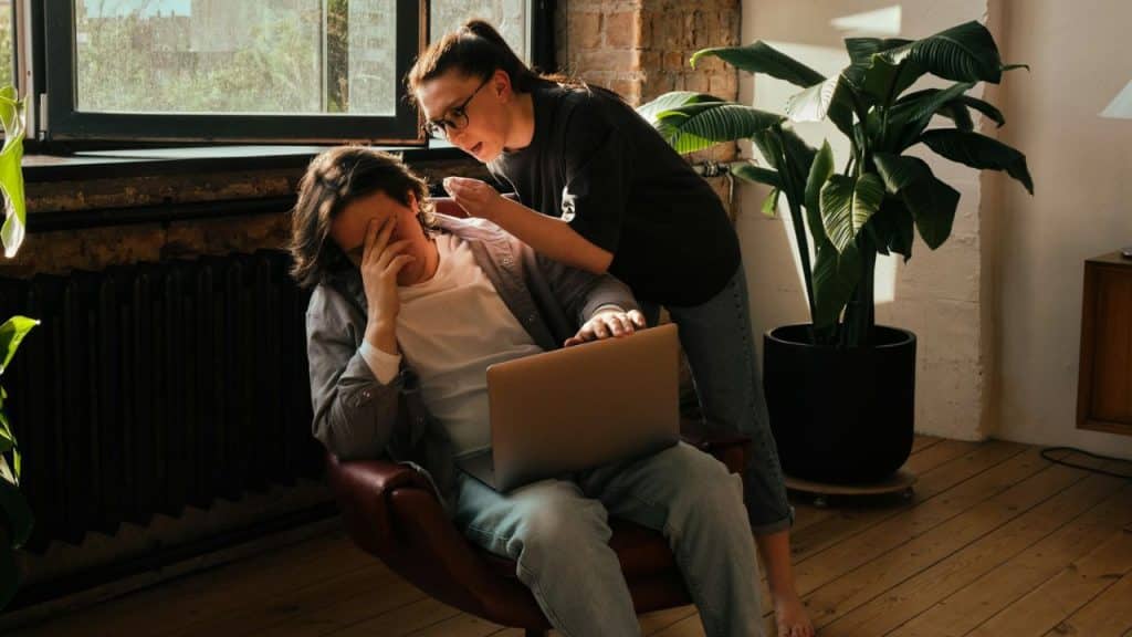 A distressed person is sitting in a chair with a laptop and their hand covering their face, while another person is standing and talking to them.